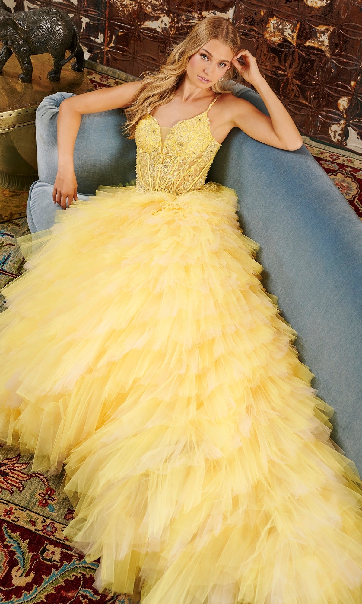 A woman in the Tiffany Tiered Ruffled Tulle Prom Ball Gown 16115, featuring a bright yellow hue and sheer corset bodice, sits on a blue sofa with her arm resting on the back, in a room decorated with a patterned rug and animal figurine.